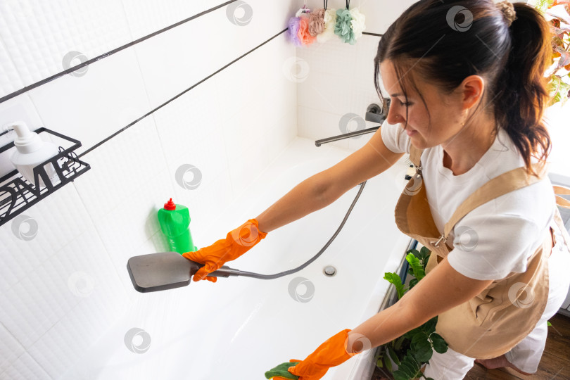 Скачать Женщина в резиновых перчатках и фартуке вручную моет ванну в ванной комнате дома с помощью губки для мытья посуды и чистящего средства в бутылочке. Наведение порядка, чистоты и гигиены в доме, услуги по уборке фотосток Ozero