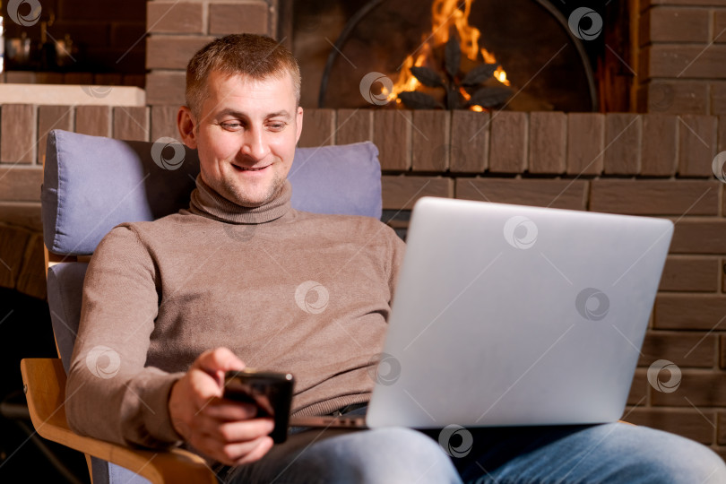 Скачать Мужчина сидит в кресле с ноутбуком и сотовым телефоном в руках фотосток Ozero