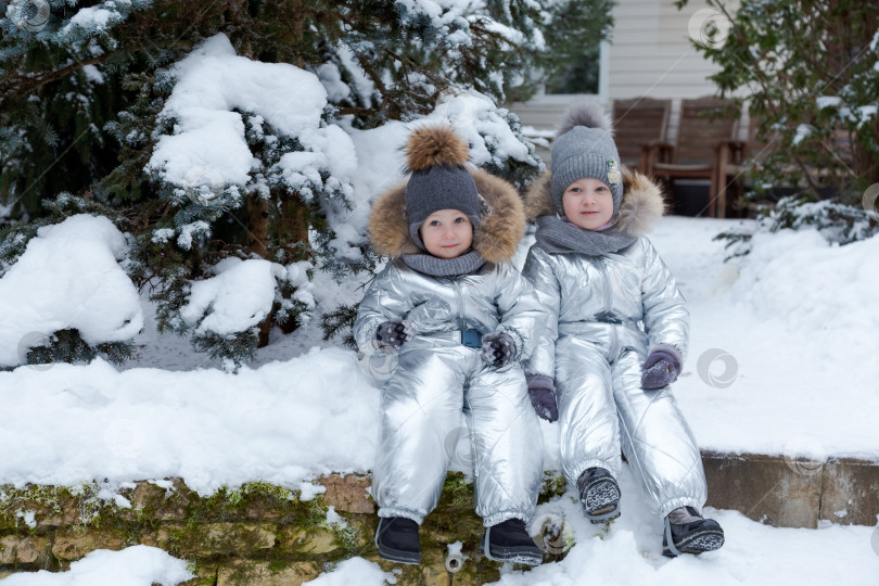 Скачать Двое детей сидят на сугробе в серебристых снежных костюмах фотосток Ozero