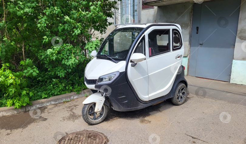 Скачать Рыбинск, Россия, 10.06.2025 В яркий солнечный день на тротуаре перед зданием припаркован белый трехколесный электрический велосипед с кабиной. фотосток Ozero