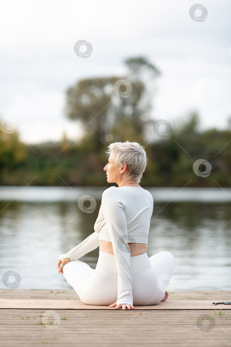 Скачать Взрослая женщина в белом костюме занимается йогой на берегу реки фотосток Ozero