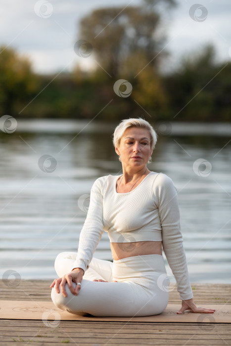 Скачать Взрослая женщина в белом костюме занимается йогой на берегу реки фотосток Ozero