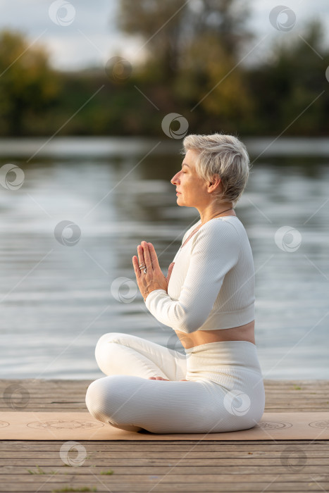Скачать Взрослая женщина в белом костюме занимается йогой на берегу реки фотосток Ozero