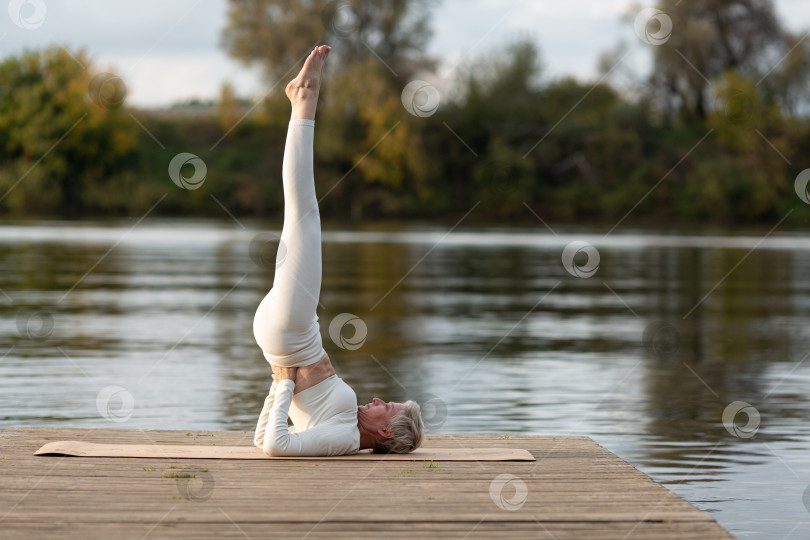 Скачать Взрослая женщина в белом костюме занимается йогой на берегу реки фотосток Ozero