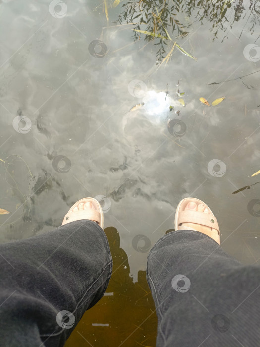 Скачать Ноги человека в сандалиях со спокойным и капризным чувством свисают над водой, олицетворяя расслабление и созерцание на фоне отражения воды. фотосток Ozero