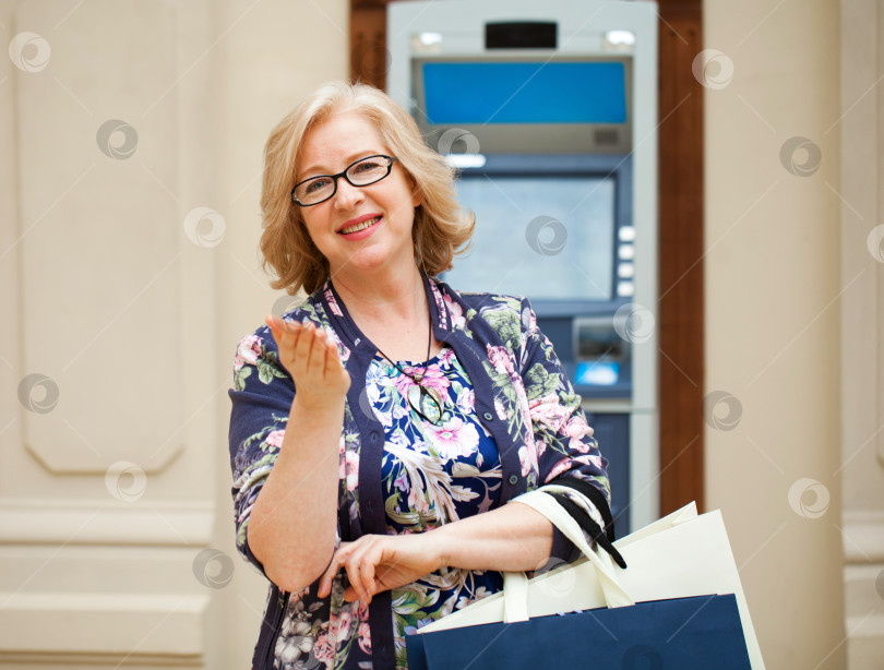 Скачать Пожилая женщина с покупкой блондинки стоит на заднем плане у банкомата торгового центра фотосток Ozero