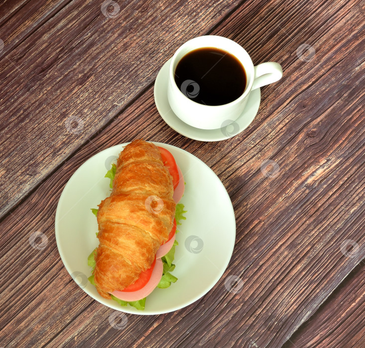 Скачать Чашка черного кофе на блюдце и бутерброд из круассана с колбасой, помидорами и листьями салата на деревянном столе. фотосток Ozero