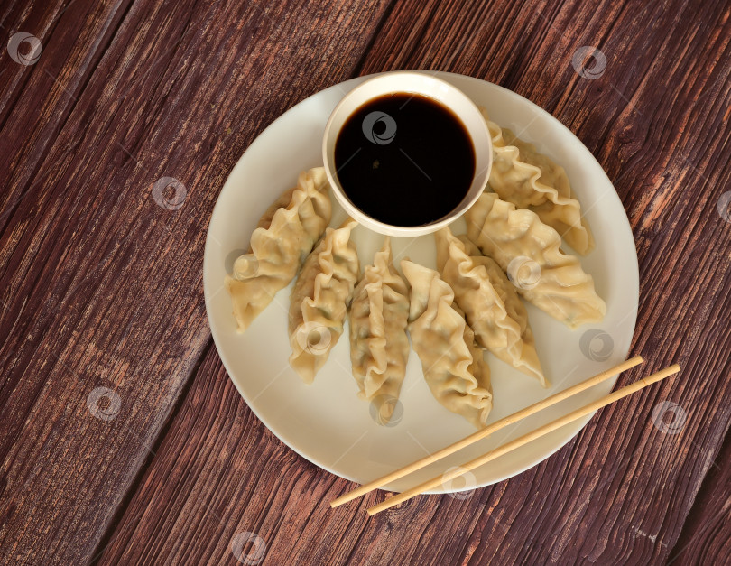 Скачать Свежеприготовленные китайские вонтоны в круглой тарелке, чашка с соусом и деревянные палочки для еды на деревянном столе. фотосток Ozero