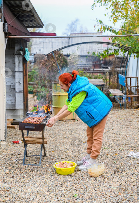 Скачать Прохладным осенним днем в России 50-летняя русская женщина в сельской местности готовит маринованную курицу на гриле на открытом воздухе, держа шашлык на открытом огне. фотосток Ozero