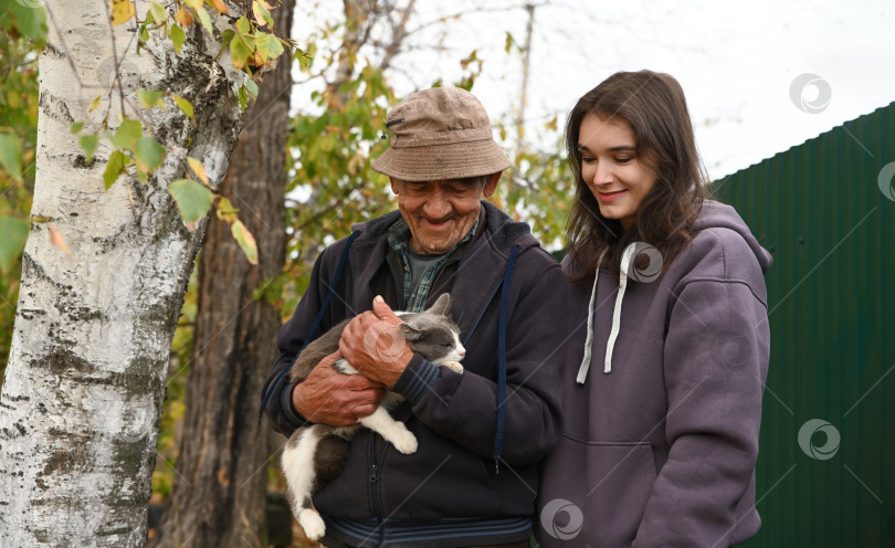 Скачать Пожилой русский дедушка и его взрослая внучка смотрят на серую кошку на улице в деревенском саду. Семейная связь в сельской осени. фотосток Ozero