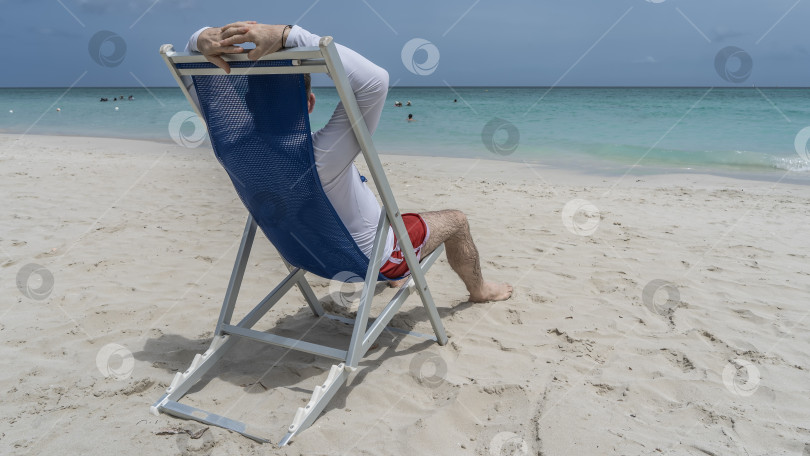 Скачать Мужчина сидит в шезлонге на песчаном пляже и смотрит на океан. фотосток Ozero