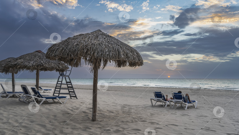 Скачать Закат на берегу океана. Двое людей лежат на шезлонгах фотосток Ozero