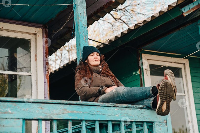 Скачать Русская женщина отдыхает у себя во дворе, наслаждаясь природой, прохладным воздухом и мирным уединением осенним или весенним днем, ощущая комфорт и спокойно мечтая. фотосток Ozero