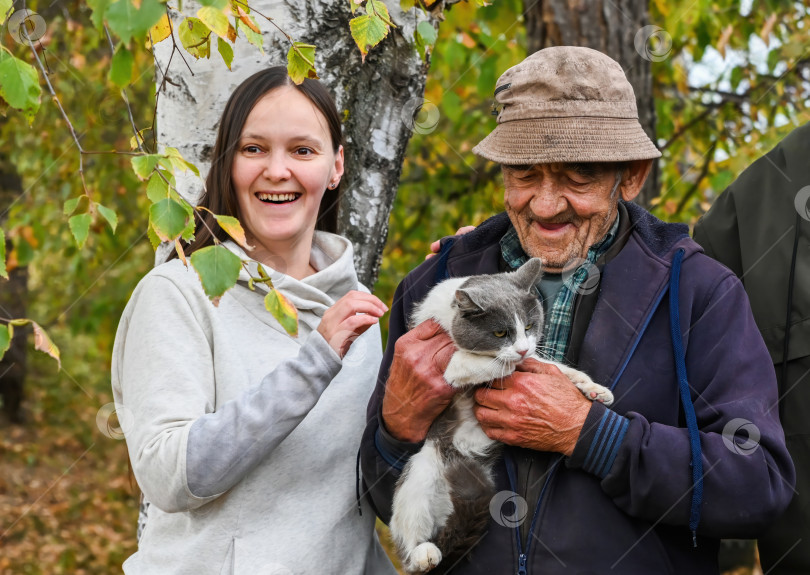 Скачать Улыбающаяся молодая женщина и ее пожилой дедушка держат серо-белого кота на улице осенним днем, в окружении берез и желтых листьев. фотосток Ozero