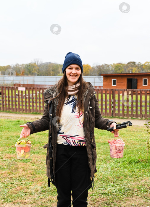 Скачать Улыбающаяся русская туристка, держащая в руках ведра с кормом для животных в загородном парке осенним днем. Момент из жизни в загородном путешествии на свежем воздухе. фотосток Ozero