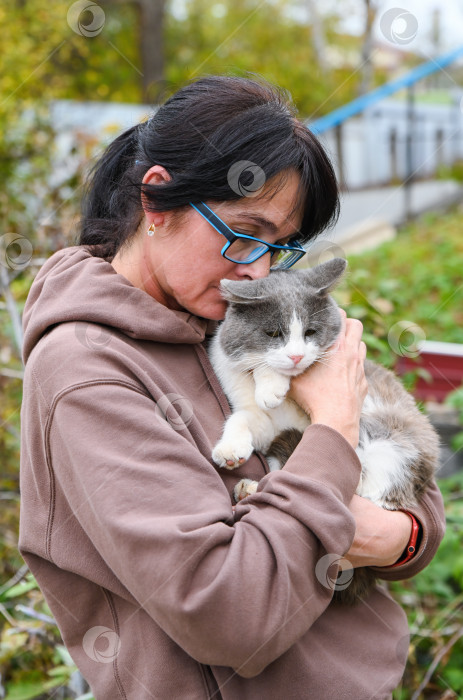 Скачать Русская женщина в коричневой толстовке с капюшоном нежно целует свою серо-белую кошку на открытом воздухе в осеннем саду, демонстрируя любовь и теплоту деревенской жизни. фотосток Ozero