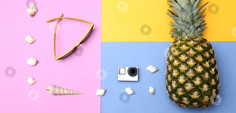 Скачать Ананас, солнцезащитные очки, соломенная шляпа, экшн-камера и ракушки на желто-сине-розовом фоне. Концепция тропического летнего отдыха с ананасом. фотосток Ozero