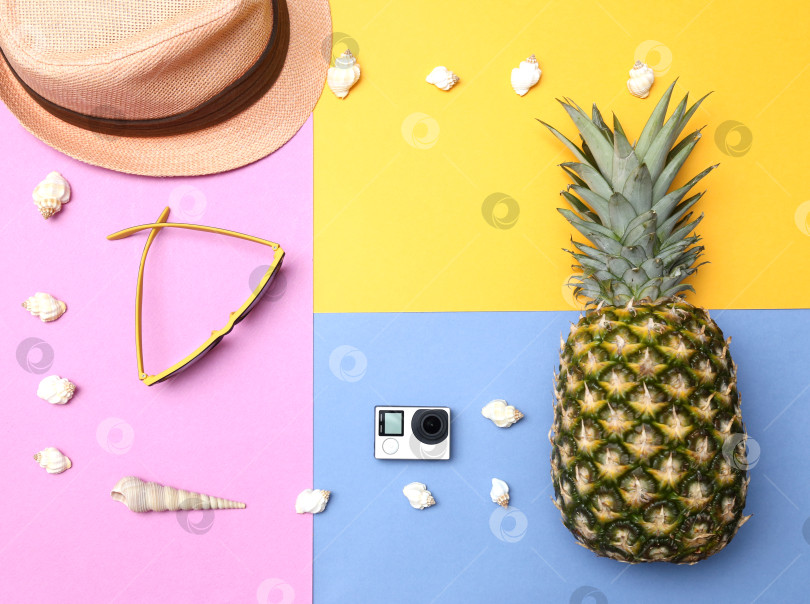 Скачать Ананас, солнцезащитные очки, соломенная шляпа, экшн-камера и ракушки на желто-сине-розовом фоне. Концепция тропического летнего отдыха с ананасом. фотосток Ozero