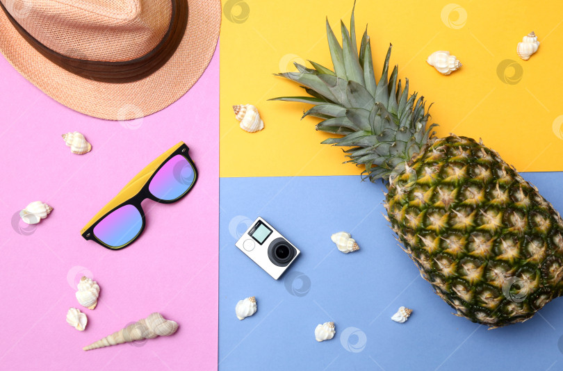 Скачать Ананас, солнцезащитные очки, соломенная шляпа, экшн-камера и ракушки на желто-сине-розовом фоне. Концепция тропического летнего отдыха с ананасом. фотосток Ozero
