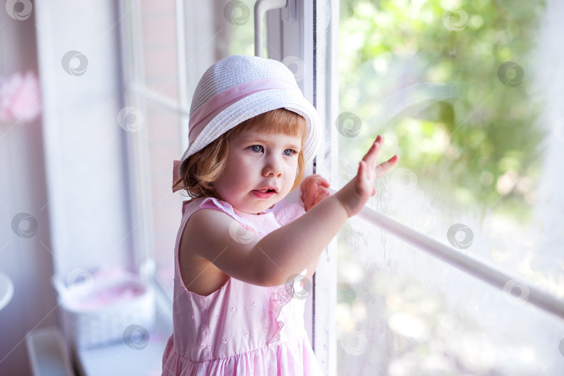 Скачать Грустная малышка в розовом платье и шляпке смотрит в большое окно фотосток Ozero