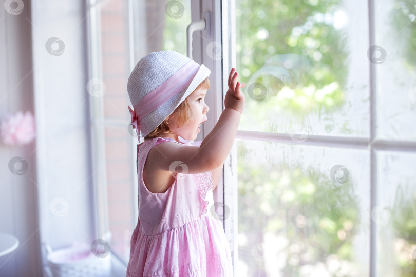 Скачать Маленькая девочка в розовом платьице и шляпке смотрит в большое окно фотосток Ozero