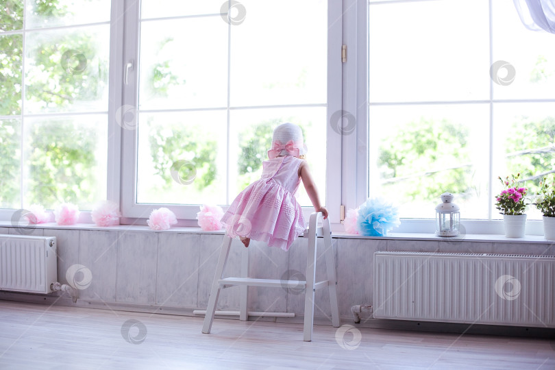 Скачать Маленькая девочка в розовом платьице и шляпке смотрит в большое окно фотосток Ozero