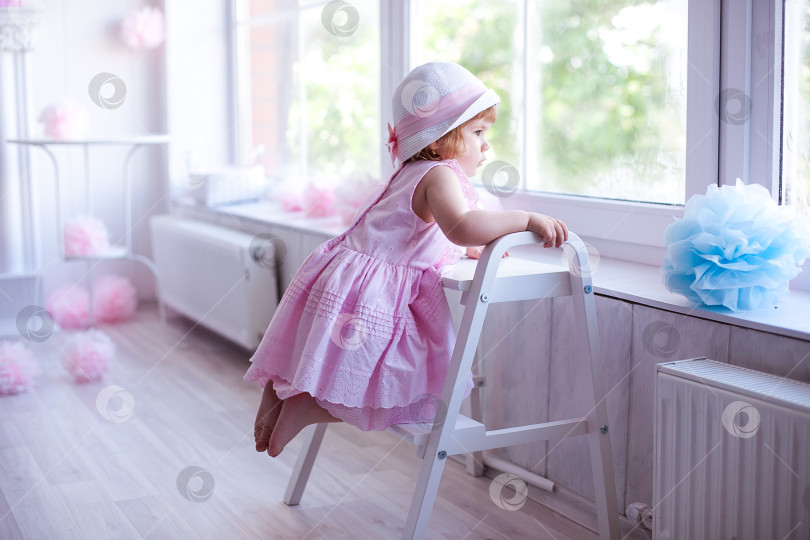 Скачать Грустная малышка в розовом платье и шляпке смотрит в большое окно фотосток Ozero