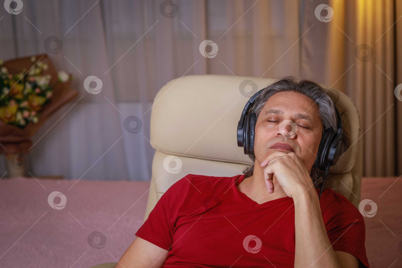 Скачать 50-летний мужчина слушает музыку в наушниках дома, сидя в кресле. фотосток Ozero