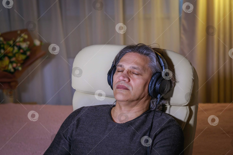 Скачать 50-летний мужчина слушает музыку в наушниках дома, сидя в кресле. фотосток Ozero