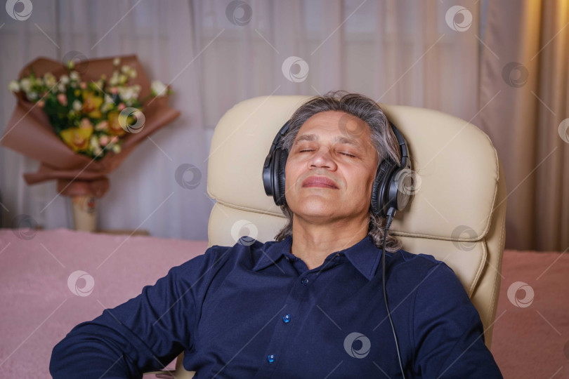 Скачать 50-летний мужчина слушает музыку в наушниках дома, сидя в кресле. фотосток Ozero