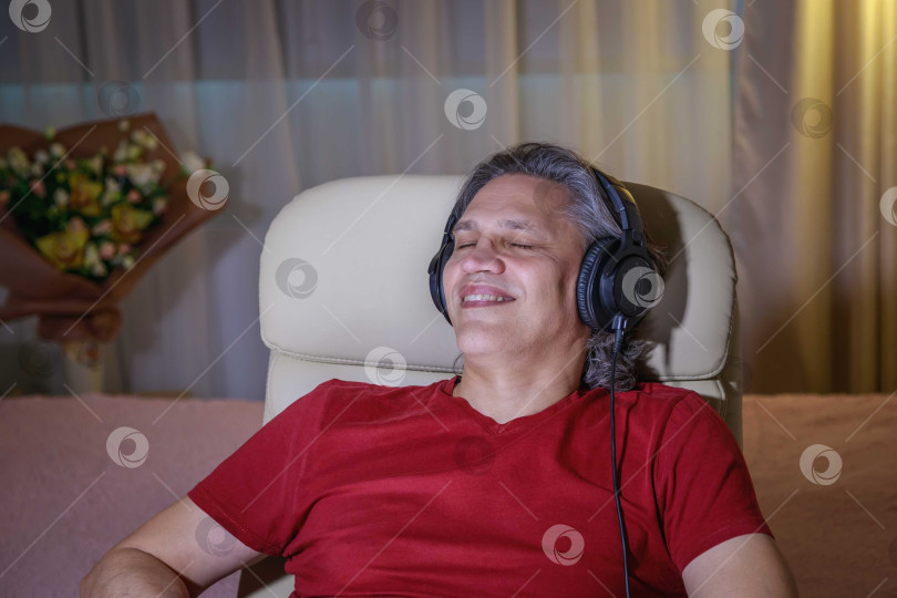 Скачать 50-летний мужчина слушает музыку в наушниках дома, сидя в кресле. фотосток Ozero