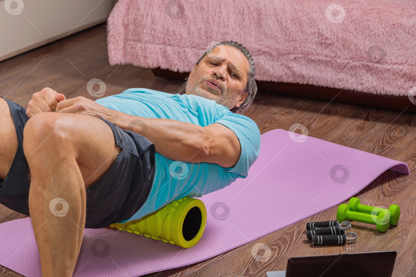 Скачать 50-летний мужчина выполняет упражнения, лежа дома на коврике и глядя в компьютер фотосток Ozero