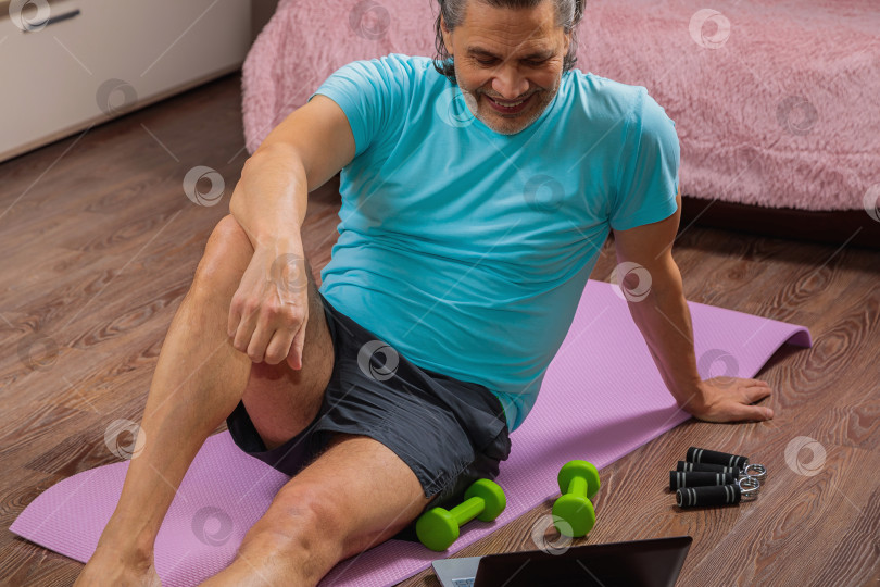 Скачать 50-летний мужчина выполняет упражнения, лежа дома на коврике и глядя в компьютер фотосток Ozero