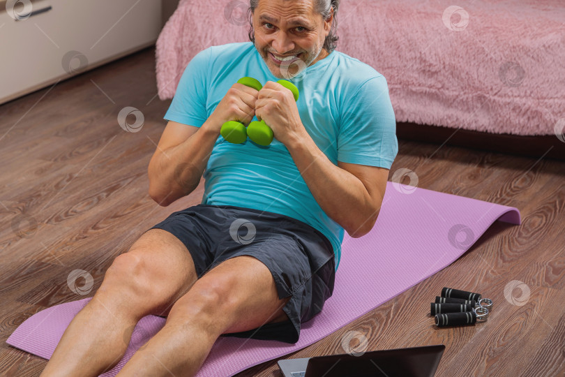 Скачать 50-летний мужчина выполняет упражнения, лежа дома на коврике и глядя в компьютер фотосток Ozero