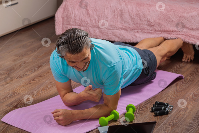 Скачать 50-летний мужчина выполняет упражнения, лежа дома на коврике и глядя в компьютер фотосток Ozero