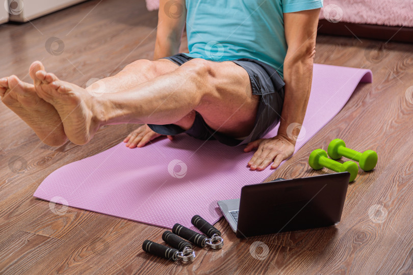Скачать 50-летний мужчина выполняет упражнения, лежа дома на коврике и глядя в компьютер фотосток Ozero