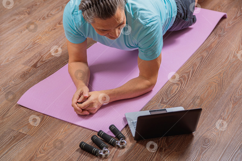 Скачать 50-летний мужчина выполняет упражнения, лежа дома на коврике и глядя в компьютер фотосток Ozero