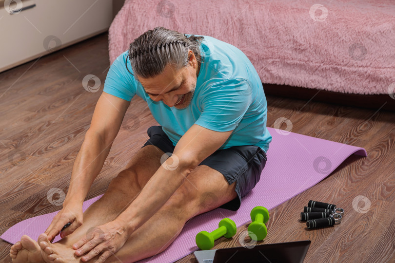 Скачать 50-летний мужчина выполняет упражнения, лежа дома на коврике и глядя в компьютер фотосток Ozero
