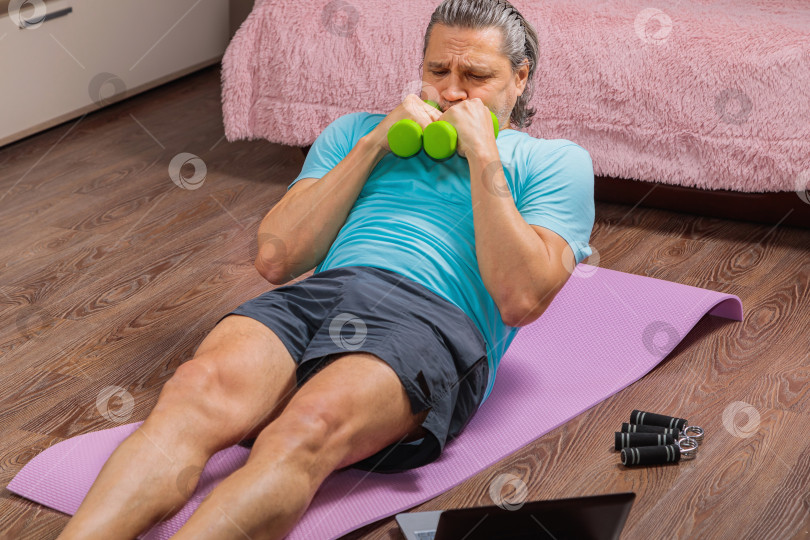 Скачать 50-летний мужчина выполняет упражнения, лежа дома на коврике и глядя в компьютер фотосток Ozero