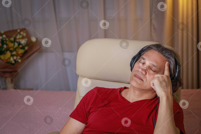 Скачать 50-летний мужчина слушает музыку в наушниках дома, сидя в кресле. фотосток Ozero