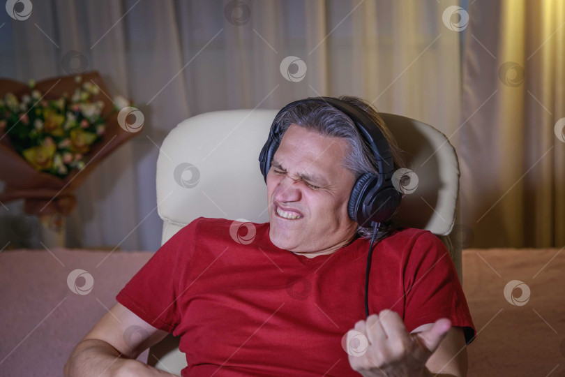 Скачать 50-летний мужчина слушает музыку в наушниках дома, сидя в кресле. фотосток Ozero