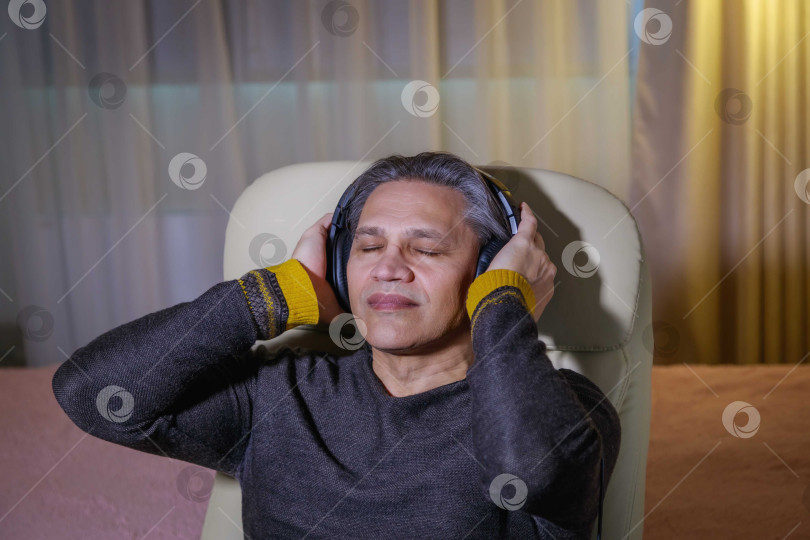 Скачать 50-летний мужчина слушает музыку в наушниках дома, сидя в кресле. фотосток Ozero