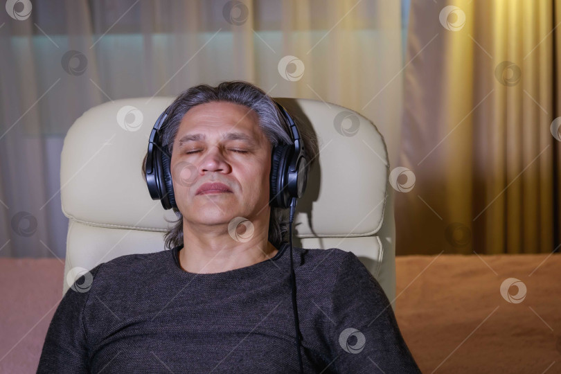 Скачать 50-летний мужчина слушает музыку в наушниках дома, сидя в кресле. фотосток Ozero