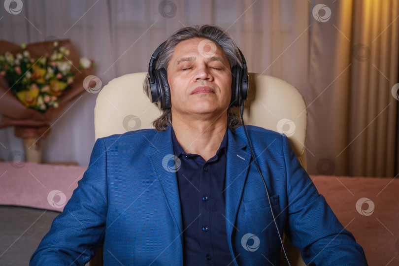 Скачать 50-летний мужчина слушает музыку в наушниках дома, сидя в кресле. фотосток Ozero