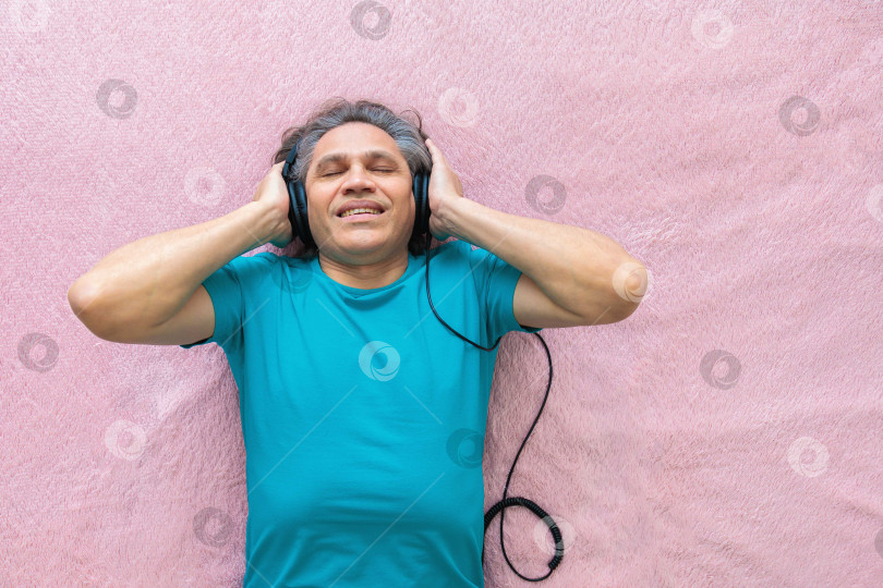Скачать 50-летний мужчина слушает музыку в наушниках дома, лежа на кровати фотосток Ozero