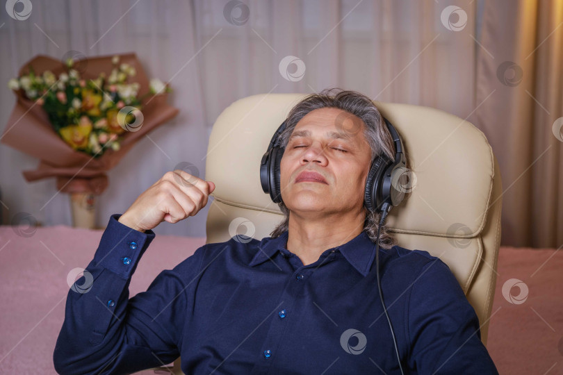 Скачать 50-летний мужчина слушает музыку в наушниках дома, сидя в кресле. фотосток Ozero