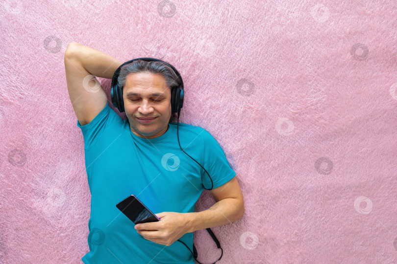 Скачать 50-летний мужчина слушает музыку в наушниках дома, лежа на кровати фотосток Ozero