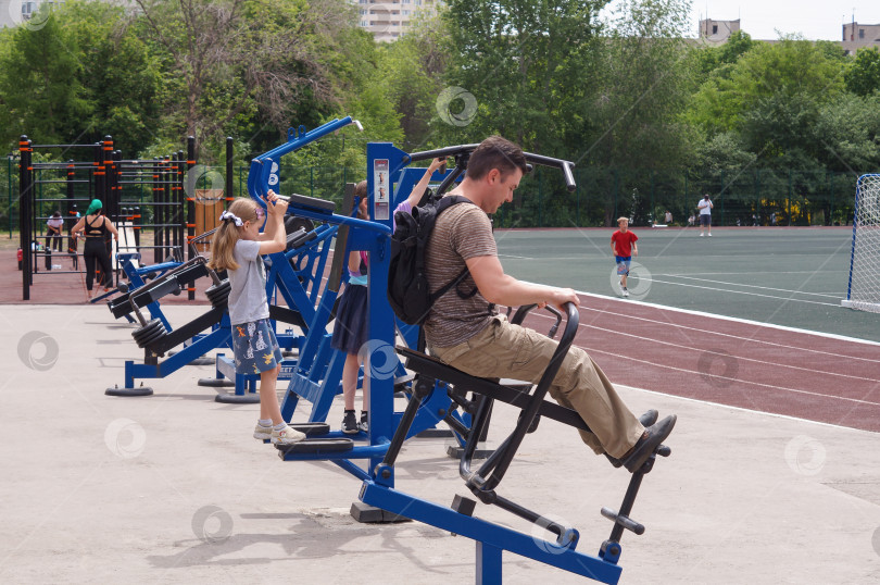 Скачать Саратов, Россия - 01 июня 2024 года. Мужчина занимается силовой тренировкой на тренажере в парке на спортивной площадке вместе с детьми фотосток Ozero