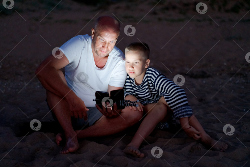 Скачать Мужчина и мальчик сидят на пляже и смотрят в сотовый телефон фотосток Ozero