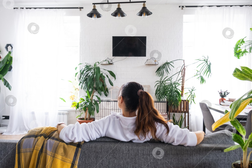 Скачать Женщина дома в белом интерьере сидит на диване и смотрит телевизор, макет черного экрана. Домашний отдых, досуг фотосток Ozero
